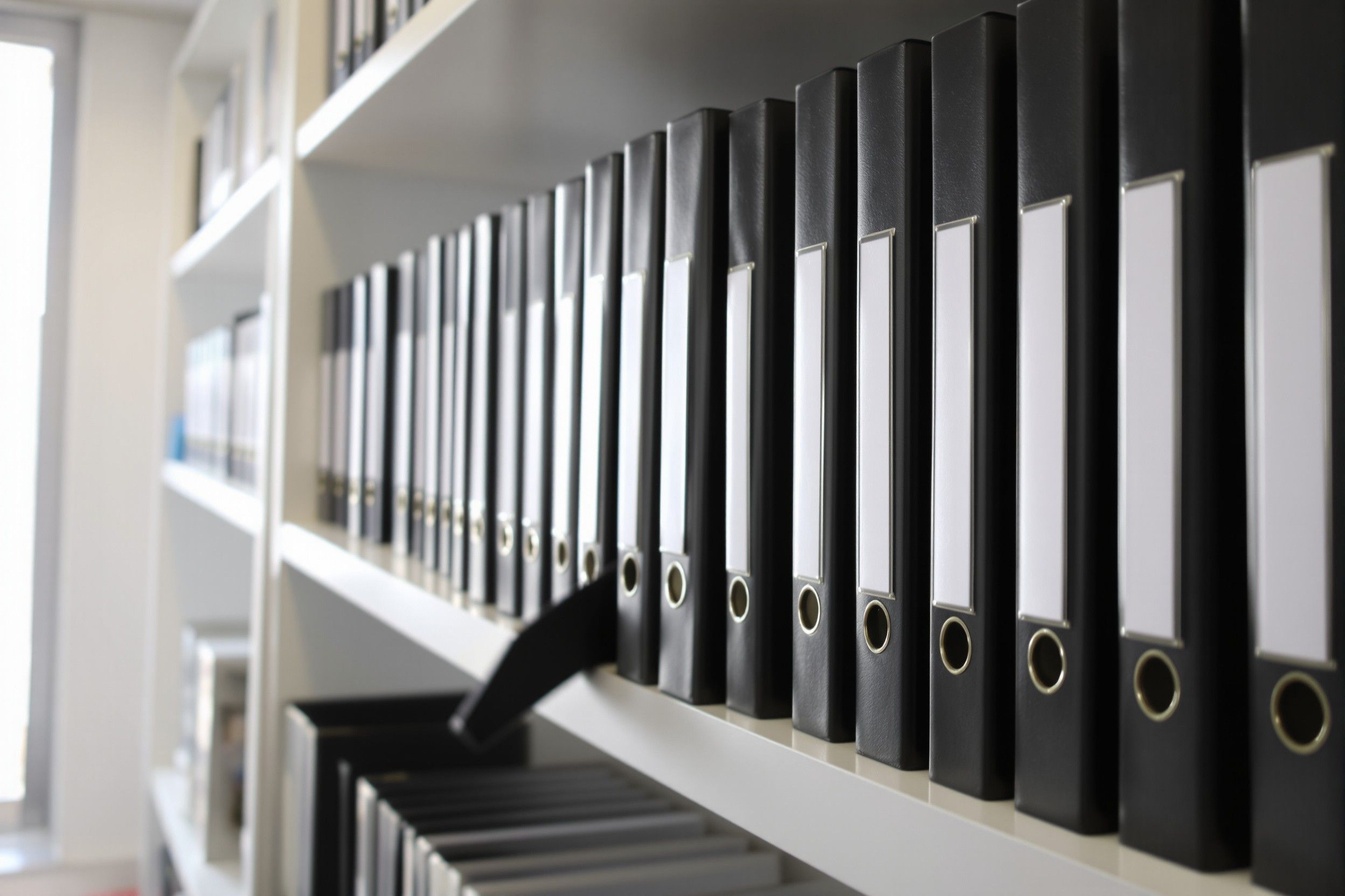 A row of black ring binders with blank spine labels on a white shelf